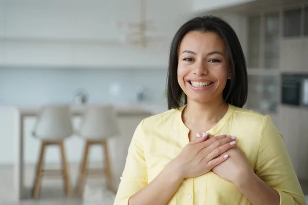 Grateful smiling woman hold hands on chest express thankfulness, love, standing at home. Touched happy female feels gratitude, thanks, appreciates help, good deed, charity and donation.