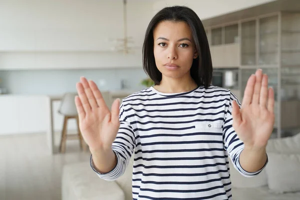 Young woman showing stop gesture by palms, warning sign of prohibition or rejection, looking at camera at home. Serious strict female protesting against domestic violence, abuse, gender discrimination