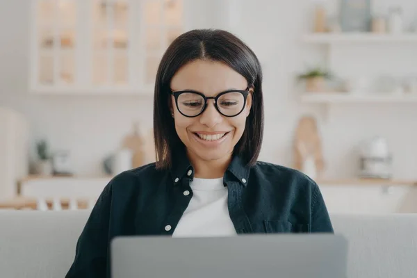 Smiling woman in glasses working at laptop, learning online at home. Portrait of young female looking at computer screen, chatting online in social network, making video call, watching series.