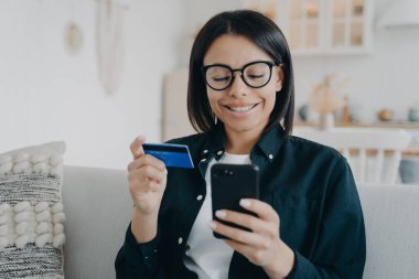 Smiling female holding bank credit card and smartphone. Young woman pays for purchases in online store using banking service, shopping, making secure payment, sitting on sofa at home. E-commerce.