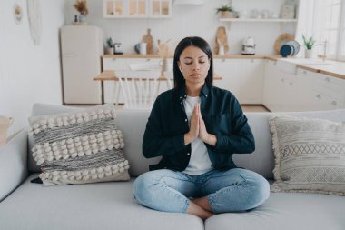 Young mixed race woman is practicing yoga at home. Girl sitting on couch in lotus pose and meditating with her eyes closed. Tranquility and stress relief. Peaceful mind, zen and wellness.