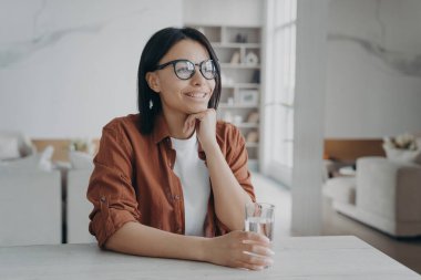 Smiling young woman enjoy purified mineral water holding glass sitting at kitchen at home. Happy female wearing glasses with pure water drink at table. Healthy lifestyle, diet concept.