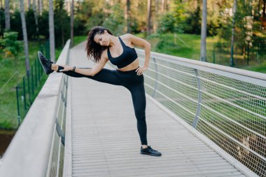 Determined sportswoman stretches legs on bridge, wears active wear, demonstrates good flexibility, poses outdoor against forest background. Woman runner prepares for jogging, has summer workout