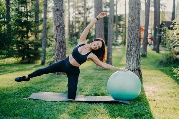 Horizontal shot of active slim woman with cheerful expression leans aside does exercises with fitness ball, dressed in sportsclothes, poses outdoor against tall trees on karemat. Healthy lifestyle