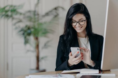 Güzel, gülümseyen kadın serbest çalışan, akıllı telefona odaklanmış, gelen mesaja geri bildirim yollayan, birlikte çalışma alanında oturan, resmi giyinen, bilgisayarda çalışan, internette sörf yapan,