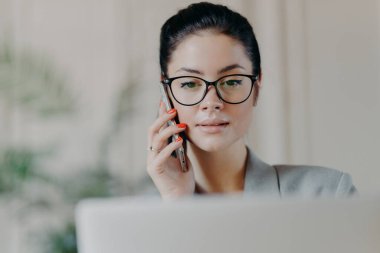 Koyu renk saçlı, ciddi ifadesi, makyajı olan, cep telefonuyla konuşan, ekrana odaklanmış, iş ortağını arayan, iş ile ilgili konuları konuşmak isteyen, ofis içi konuları olan deneyimli bir kadının yatay çekimi.