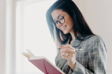 Hoş görünümlü esmer kadının yan fotoğrafları mutlu bir ifadeye sahiptir. Planlama yapar, uzaktan iş yapar, planlama yapar, optik gözlük takar ve resmi kıyafetler giyer, pencerenin yanında poz verir.