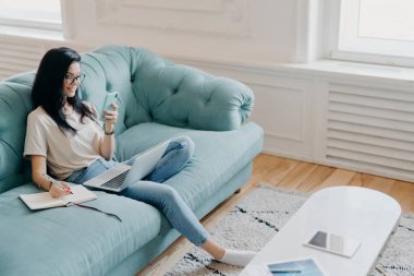 Samrtphone ekranına odaklanmış güzel esmer bir kadının resmi. Gerekli bilgileri not defterine yazıyor. Webinar eğitimi izliyor, rahat koltukta rahat bir odada poz veriyor.