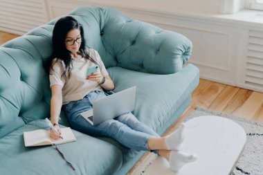 İnsanlar, uzaktan öğrenme, teknolojiler, serbest çalışma kavramı. Ciddi esmer kadın günlüğe bilgi yazıyor, cep telefonu tutuyor, uzaktan kumandalı işe odaklanmış, dizüstü bilgisayarla çalışıyor.