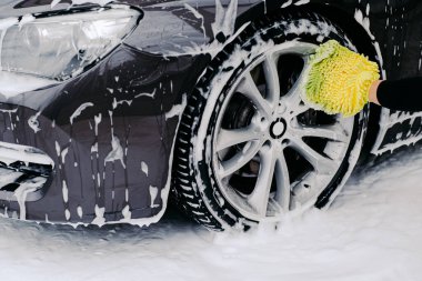 Cropped shot of black automobile washed by service worker at carwash. Vehicle covered with foam bubbles. Focus on wheel.