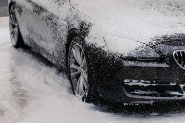 Horizontal shot of black automobile covered with bubble soap foam at carwash. Detail cleaning. Purifying concept