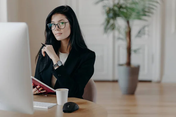 Photo of serious woman writes information in notebook, studies new online business course, poses in coworking space with computer, works on project, writes some ideas. Business and work concept