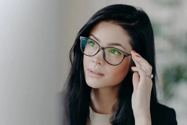 Close up shot of brunette serious lady keeps hand on rim of spectacles, concentrated at computer screen, watches online video, poses indoor, browses web page, creats project, poses in office