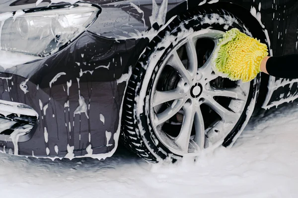Cropped shot of black automobile washed by service worker at carwash. Vehicle covered with foam bubbles. Focus on wheel.