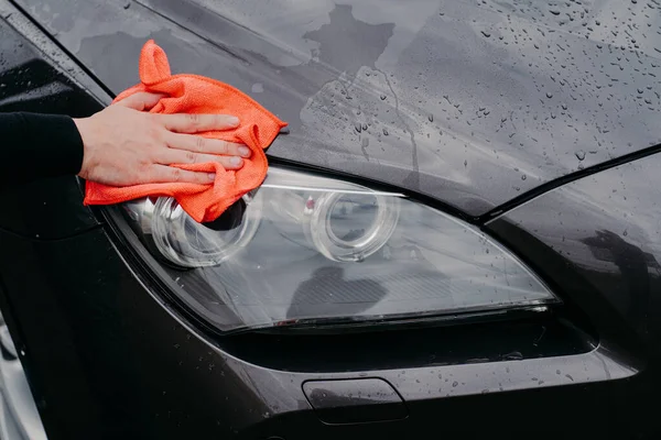 Mans hand with rag wiping cars headlamp. Wet black automobile cleanes at car wash. Auto cleaning and service concept