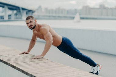 Fit muscular man does plank push up exercise leads healthy lifestyle wears sport trousers and sneakers poses outdoor near bridge. Motivated sportsman pushing hard. Sport, motivation and determination