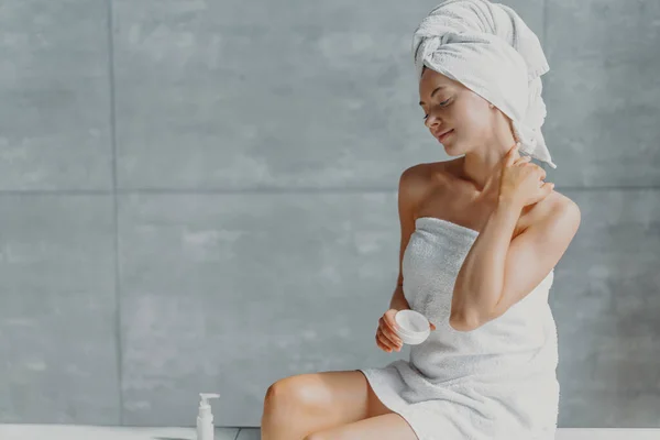 Satisfied spa woman sits in profile applies body cream for perfect skin, enjoys pampering and beauty treatment poses wrapped in towel against grey wall in bathroom. Taking good care of own body