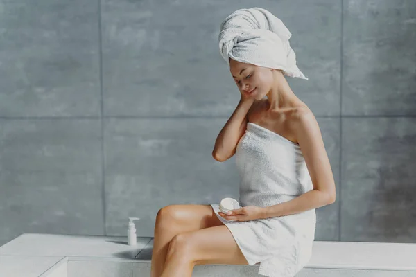 Refreshed young European female applies anti wrinkle cream, poses in bathroom, wrapped in bath towels, prevents signs of skin aging, has clean body after showering. Wellness, wellbeing concept