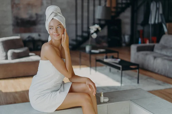 Women, cosmetology, beauty and hygiene concept. Relaxed thoughtful woman wrapped in bath towel, applies anti wrinkle cream or body lotion, poses in living room at home, looks pensively right