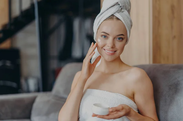 健康な滑らかな肌を持つかなり若い女性は 顔クリームを適用します シャワーを浴びた後 頭の上にラップタオルを身に着けています 快適なソファの上にポーズ 衛生手順 美容と美しさの概念 — ストック写真