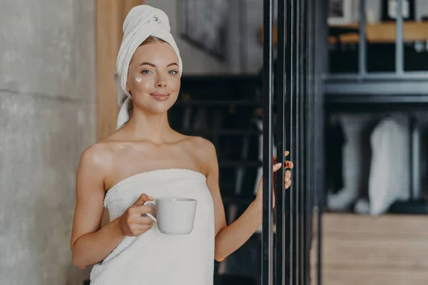 Photo of satisfied young female model wears cream on face, stands satisfied after taking bath, wrapped in white soft towel, drinks coffee or tea, poses over home interior. Anti aging treatment