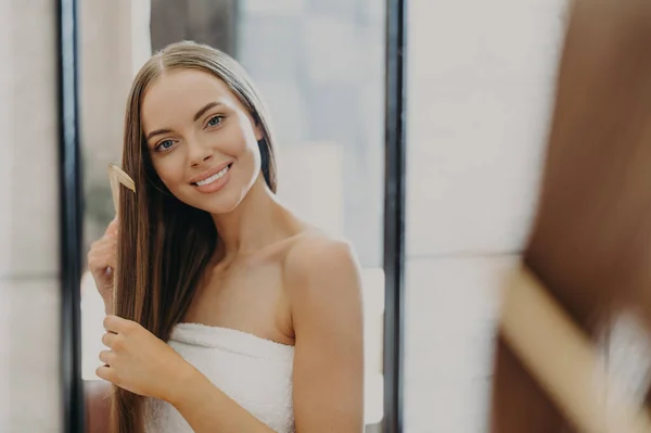 Portrait of beautiful gorgeous woman with bare shoulders combes her straight hair, smiles broadly and looks at her reflection in mirror, poses in bathroom, wrapped in towel. Women beauty concept
