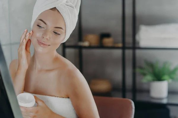 健康可爱的女人用保湿霜 裹在浴巾里 细心地看着镜子里的倒影 沐浴后享受肌肤护理治疗 在浴室里摆姿势 美的概念 — 图库照片
