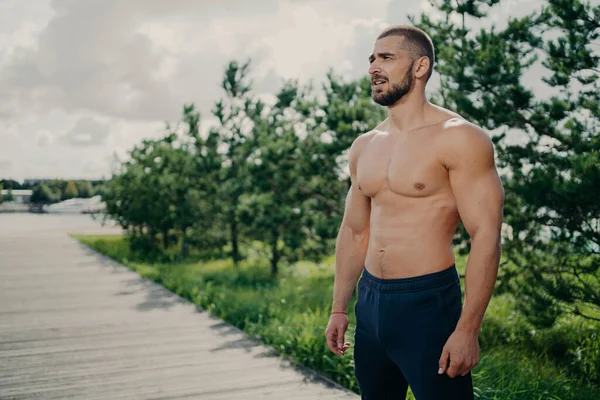 Confident sporty man bodybuilder has workout outdoor, looks thoughtfully into distance, wears shorts, has muscular body, strong biceps, stands over trees and sky background, shows strong torso