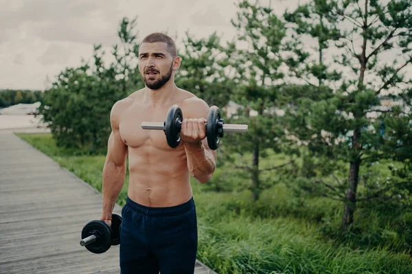 Body care and power lifting concept. Strong muscular athlete bearded man raises dumbbells, lifts heavy barbells, performs shoulder press, poses outside against nature background. Sport exercising