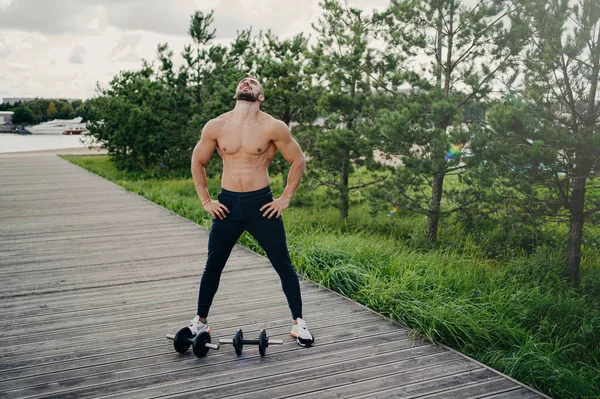 健康な筋肉の男は裸の胴で立ち 頭上に頭を上げ 深く呼吸し スポーツ服を着て バーベルでワークアウトをし 屋外でポーズを取ります ボディビル アクティブライフスタイル フィットネスコンセプト — ストック写真