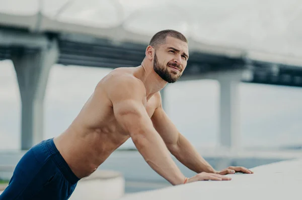 スポーツの動機付けられた目的意識のあるひげそりのスポーツの裸の胸の男の側面図は プッシュアップ運動を実行し 努力が橋の上に傾き したがって距離を見て 筋肉の体を持っている 外で動作します — ストック写真