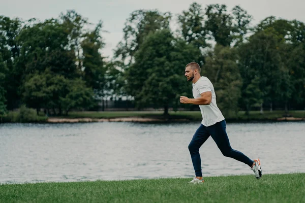 Handsome athlete bearded man runs outdoors in morning poses near river enjoys nature and fresh air, demonstrates endurance and motivation, has morning workout every day, stays fit healthy strong