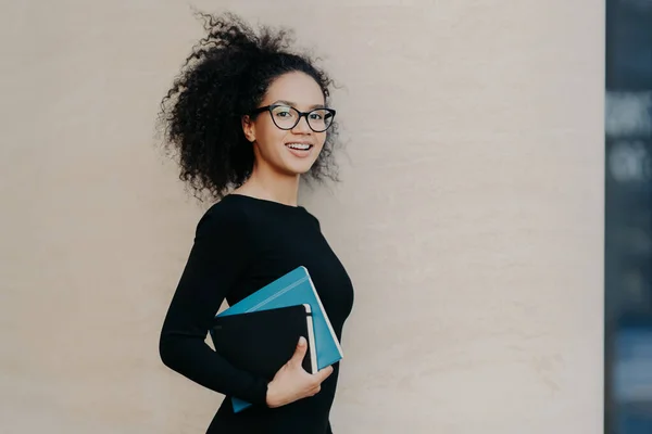 Sideways shot of beautiful dark skinned curly college student dressed in black casual outfit, carries textbook, notepad, smiles positively, isolated on grey background. People and studying concept