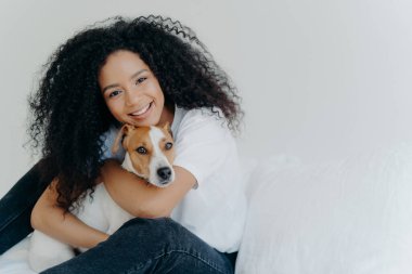 Afro-Kircut 'lu çekici genç bir kadının fotoğrafı, aşk köpeğiyle kucaklaşması, evcil hayvanla ilgilenmesi, nazikçe gülümsemesi, günlük kıyafetler giymesi, beyaz arka planda izole edilmesi, yatağa oturması, tanıtımın için yer kopyalaması.