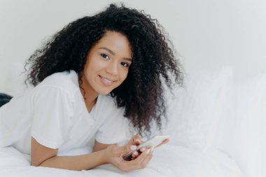 Shot of lovely curly woman with dark skin, tender smile on face, lies on white bed, wears t shirt, has relax with modern technologies, poses in bedroom, reads text message, uses free internet