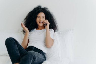 People, technology, communication and rest concept. Glad curly haired ethnic lady has telephone talk, gestures and laughs with joy, looks somewhere aside, spends time at home, sits on white bed