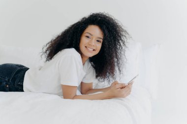 Pretty cheerful woman with Afro haircut, enjoys chatting and networking, lies on comfortable bed, looks photos, has relaxed face expression, isolated over white background. Online communication