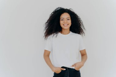Genç Afro-Amerikalı bir kadının izole edilmiş fotoğrafı beyaz tişört giyiyor, iyi duygularını ifade ediyor, evde tek başına duruyor, fotoğraf için poz veriyor, arkadaşlarıyla gündelik konuşmalar yapıyor, boş zamanlardan hoşlanıyor. İnsanlar ve mutluluk