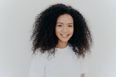 Happiness, wellbeing and positive emotions concept. Headshot of pleasant looking lady with crisp hair, smiles and laughs, has healthy skin, wears casual t shirt, models against white studio wall.
