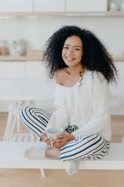 Image of lovely curly haired lady drinks coffee or tea from white mug, wears fashionable white sweater, striped pants, poses at kitchen against blurred background. People and lifestyle concept