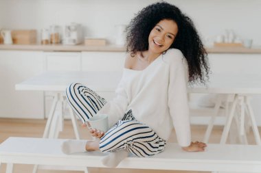 Pleased young girlfriend with Afro curly hairstyle, sits crossed legs on bench, enjoys morning coffee and domestic atmosphere, poses against kitchen background, spends time alone, has breakfast