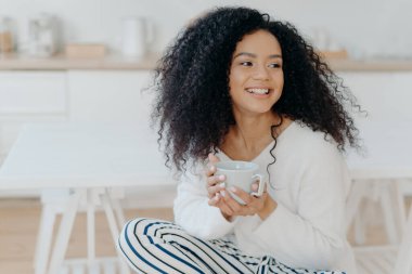 Photo of cute cheerful woman with bushy curly hair looks away with smile, holds mug of coffee, wears casual clothing, enjoys cozy calm morning, savors taste. Afro lay in kitchen during day off