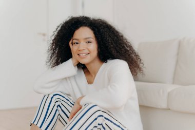 Rahatlamış, kıvırcık saçlı, kameraya nazikçe gülümseyen beyaz kazak ve çizgili pantolon giyen, modern dairenin oturma odasındaki kanepenin yanında oturan Afro-Amerikan kadının fotoğrafı.