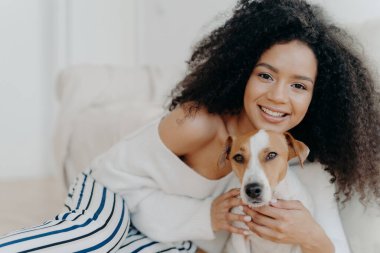 Çekici koyu tenli kadın makyajı yapıyor, kıvırcık saçları var, güzel gülümsüyor, köpeğe sarılıyor, şık kıyafetler giyiyor, yavru köpekle tatlı bir anın tadını çıkarıyor, birlikte evde olmaktan hoşlanıyor.