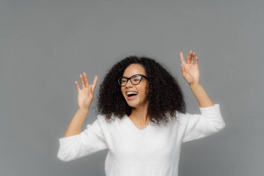 Overjoyed Afro Amerikalı kadın ellerini kaldırır, mutlu güler, hoş müzik sahiptir, optik gözlük ve beyaz rahat jumper giyer, gri arka plan üzerinde modeller, metin için yukarıdaki kopya alanı.