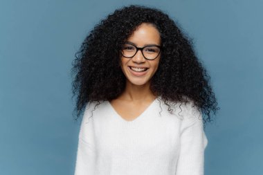 Güzel kıvırcık kadın Headshot iyi bir ruh hali içinde olmak, geniş gülümsüyor, güzel duygular ifade eder, şeffaf gözlük ve beyaz jumper giyer, mavi arka plan üzerinde izole. Afro kadın kapalı pozlar