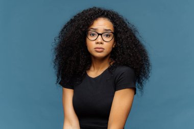 Öfkeli şaşkın Afro Amerikalı kadının fotoğraf kameraya şaşırtıcı görünüyor, rahat siyah t shirt ve gözlük giyer, mavi arka plan karşı pozlar. İnsan yüz ifadeleri ve duygu kavramı