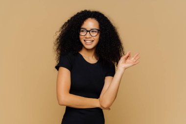 Afro saç kesimi ile Gülümseyen kadın, tamam jest yapar, mutlu bir kenara görünüyor, fareler önerisi ile kabul eder, optik gözlük ve rahat siyah t shirt giyer, iyi bir rakam, kahverengi arka plan üzerinde izole vardır
