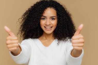 Portrait of good looking female raises thumbs, shows like gesture in camera, enjoys nice presentation, dressed in casual clothes, poses against brown background. Approval and recommendation concept