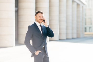 Horizontal shot of satisfied male with positive look solves banking problems while calls to operator via smart phone, wears elegant black suit, poses putdoor in urban setting. Communication concept